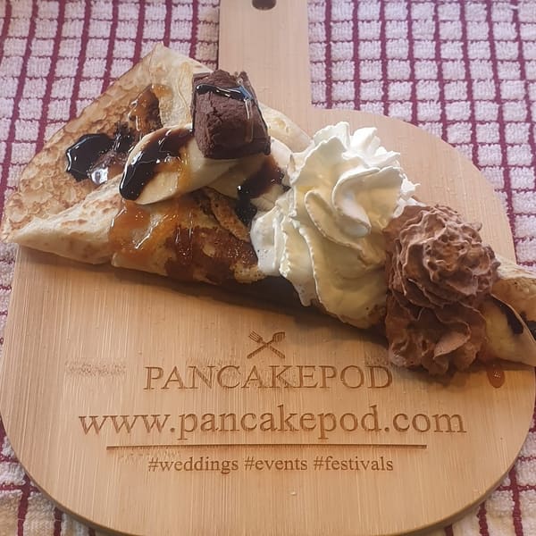 A Pancake Pod pancake folded on a wooden serving board, topped with sliced banana, chocolate sauce, caramel, a brownie piece, whipped cream and chocolate cream. The board is engraved 'Pancakepod, www.pancakepod.com, #weddings #events #festivals'